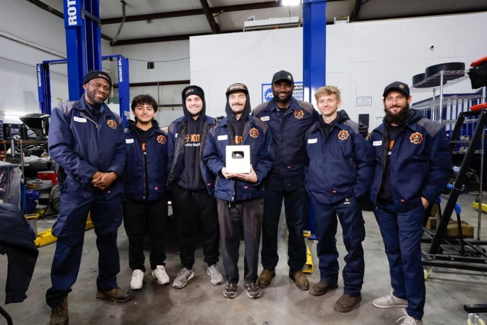 KIT'S Auto and Truck Repair Silver YouTube Creator Award in Woodbine, MD. A group of seven mechanics in matching navy blue shop uniforms posing together in an automotive repair garage, with the central person holding a Silver YouTube Creator Award.